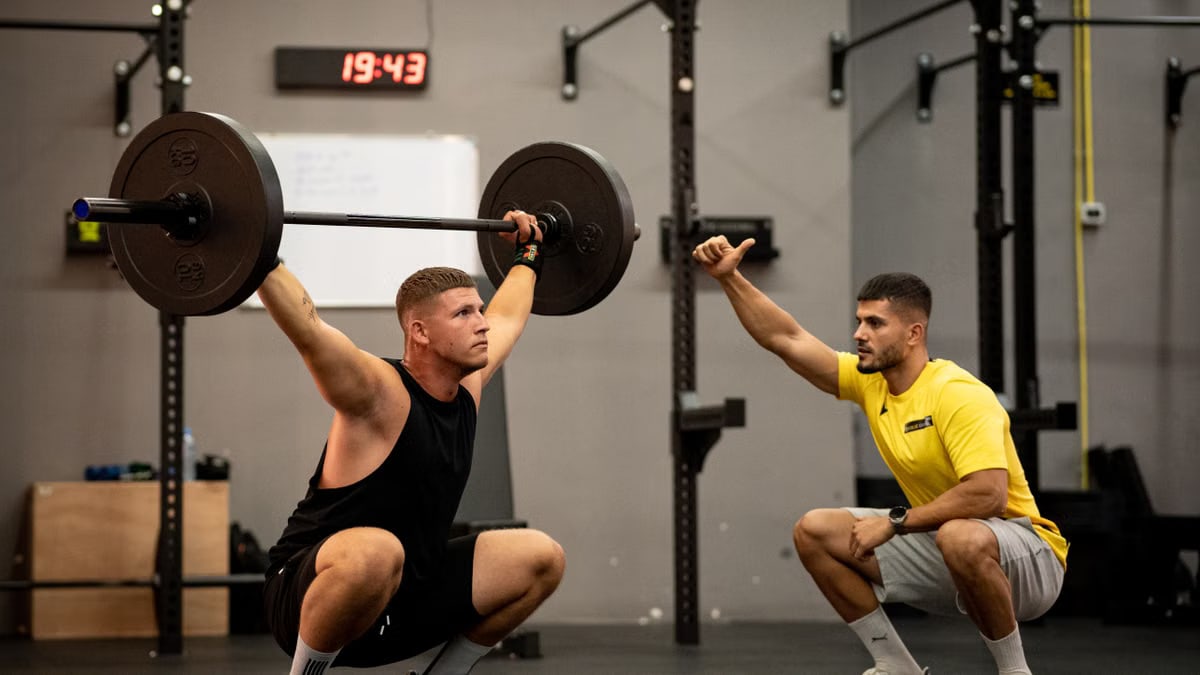 Man lifting weights under coach supervision in a gym setting, focusing on strength training.